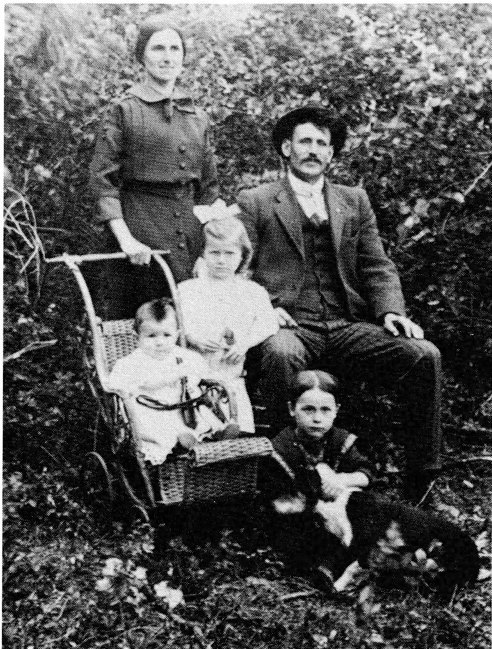 Black and white photograph of a family outdoors. A woman stands behind a man seated on a chair. A young girl stands next to the man. A baby is in a buggy to the left. A young boy sits on the ground in front.