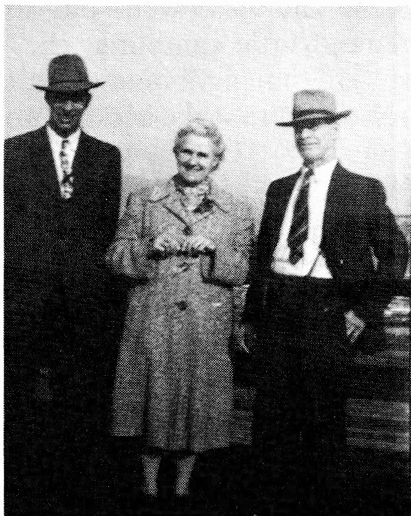 Black and white photograph of three people standing outdoors. A man in a suit and hat on the left, a woman in a dress in the middle, and another man in a suit and hat on the right.