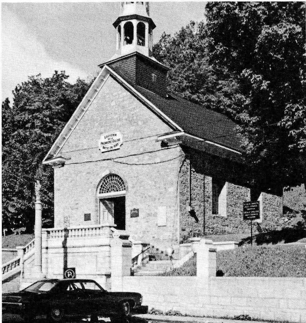 Black and white photograph of an old stone church with a small steeple and a car parked in front.