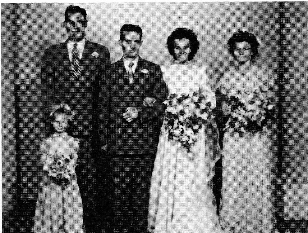 Black and white photograph of a wedding party. A bride and groom stand in the center, flanked by a best man and a bridesmaid. A young flower girl stands in front of the groom.